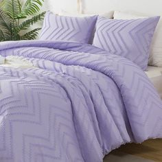 a bed with purple comforter and pillows on it in a room next to a plant