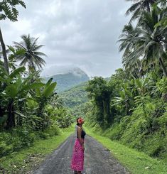 a woman in a pink dress is standing on the side of a dirt road surrounded by palm trees