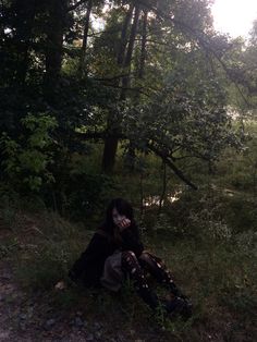 a woman sitting on the ground in front of some trees and grass with her hands to her face