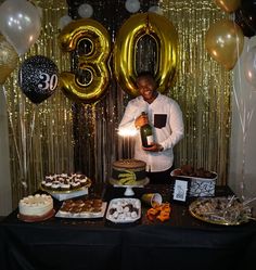 a man standing in front of a table filled with desserts and balloons, holding a bottle of booze