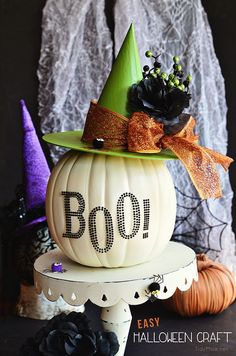 a white pumpkin with a green hat on top and boo written on the front sitting on a stand next to other halloween decorations