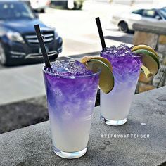 two glasses filled with purple liquid and limes on top of a stone table next to a car