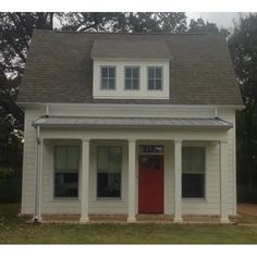 a small white house with a red door and two windows on the top floor, in front of some trees