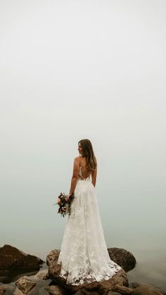 a woman in a wedding dress is standing on rocks by the water with her bouquet