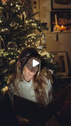 a woman sitting in front of a christmas tree looking at a tablet computer screen and smiling