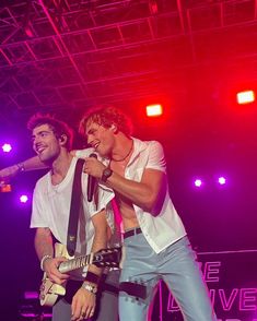 two young men singing into microphones on stage at a concert with red and purple lights