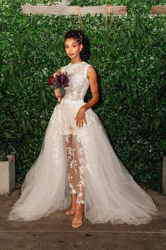 a woman standing in front of a green wall wearing a white dress and holding a bouquet