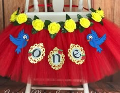 a red tutu with yellow roses and two blue birds on the top, sitting in front of a white chair