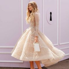 a woman wearing a dress and heels standing in front of a purple wall holding a white purse