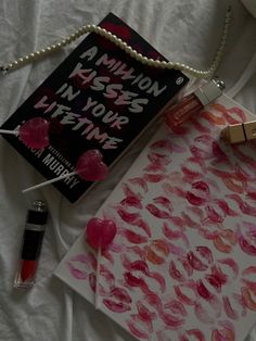 a book and lipstick are laying on a bed next to a necklace, lip balm bottle, and pen