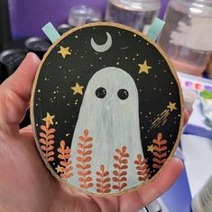 a hand holding up a painted wooden plaque with a ghost in the middle and stars on it