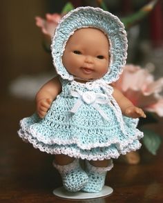 a small doll wearing a blue crocheted dress and bonnet is standing on a table