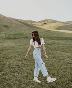 a woman walking across a lush green field with her hands in her pockets and wearing white sneakers