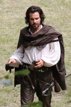 a man dressed in medieval clothing walking through the grass with his hand on his hip