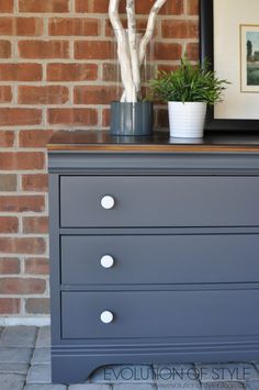 a gray dresser with two plants on top and a framed photo above it in front of a brick wall