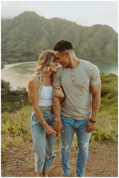 a man and woman standing next to each other on top of a hill with mountains in the background