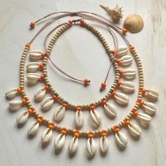 a necklace with seashells and orange beads on a white surface next to a sea shell