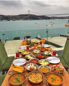 a wooden table topped with lots of plates and bowls filled with food next to a body of water