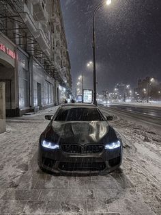 a black car parked on the side of a snow covered road at night with street lights and buildings in the background