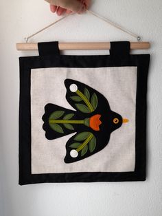 a hand is holding up a black and white wall hanging with an embroidered bird on it