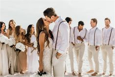 a bride and groom kissing in front of their wedding party on the beach at sunset