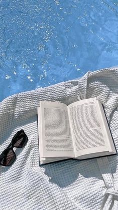 an open book sitting on top of a white towel next to a pool with sunglasses