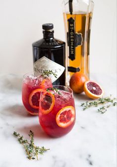 two wine glasses filled with blood oranges and garnish next to a bottle of booze