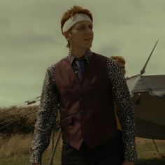 a man in a vest and tie standing next to an old fashioned boat on the grass