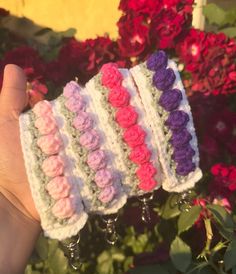 four crocheted wristbands are held up in front of some pink and purple flowers