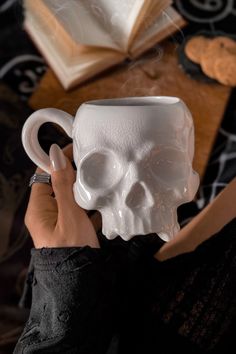 a person holding a white coffee cup with a skull on it's side, in front of an open book