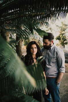 a man and woman standing next to each other in front of palm trees, looking at the camera