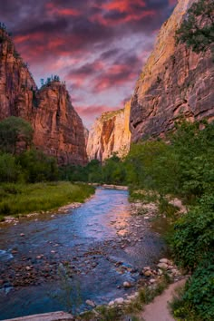 a river flowing through a lush green forest next to tall mountains under a cloudy sky