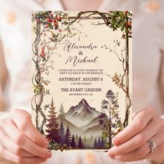 a woman holding up a wedding card with mountains and trees in the background on it