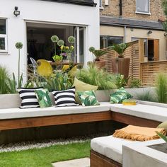 an outdoor seating area with lots of plants and pillows on the bench, in front of a house