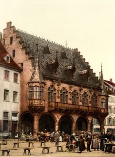 an old building with people standing around it in front of some other buildings and tables