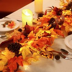 the table is set for thanksgiving dinner with autumn leaves and pine cones on it, lit by candles