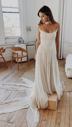 a woman in a white dress standing on a wooden floor next to a chair and window