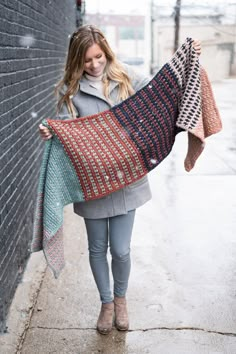 a woman is walking down the street holding up a large knitted shawl in front of a brick wall