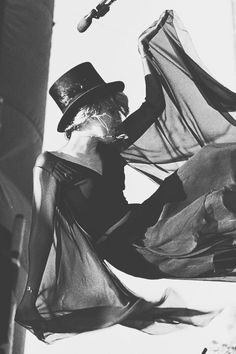 black and white photograph of woman in dress with hat holding umbrella over her head while standing on street corner