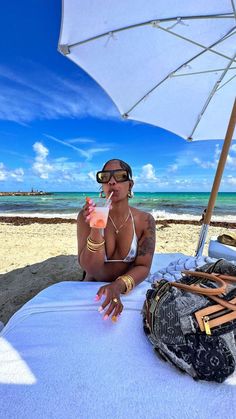 a woman sitting under an umbrella on the beach with her hand up to her mouth