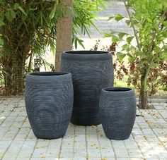 three black vases sitting next to each other on a brick floor in front of trees