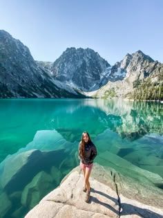 a woman standing on top of a rock next to a lake with mountains in the background