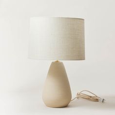 a white table lamp with a beige shade on the base and a cord plugged into it