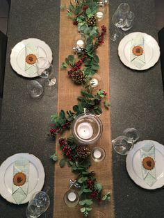 the table is set with white plates and silverware, greenery and pine cones