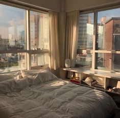 an unmade bed sitting in front of two large windows with cityscape outside