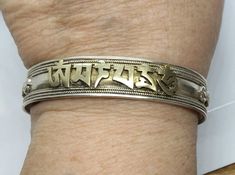 "Here's a beautiful vintage cuff bracelet with bronze overlay lettering spelling out the beautiful Buddhist Chant \"Nam Myoho Renge Kyo\" in Sanskrit. This bracelet was hand-crafted by an unidentified Native silversmith, and decorated with traditional Tibetan motifs, delicately twisted wire, and the applied Bronze letters. It's 13.5 mm wide, and measuring on the inside, this bracelet is 2 & 5/8ths inches across, about 1 & 7/8ths inches front to back, with a 1 & 1/4 inch gap between t Spiritual Bronze Cuff Bracelet As Gift, Symbolic Stamped Cuff Bracelet, Spiritual Bronze Cuff Bracelet Ideal For Gifts, Silver Spiritual Brass Bangle, Spiritual Silver-colored Brass Bangle, Spiritual Engraved Bracelets For Collectors, Spiritual Engraved Bracelets For Collectible, Spiritual Engraved Cuff Bracelet For Ceremonial Occasion, Symbolic Stamped Cuff Bangle Bracelet