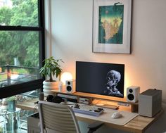 a desk with a computer, speakers and a plant in front of a large window