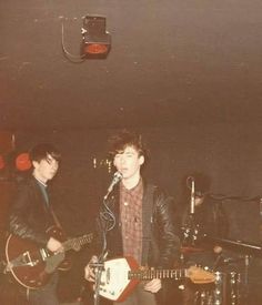 two young men are playing guitars and singing into microphones while another man stands behind them