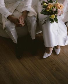 a bride and groom sitting on a chair holding each other's hands with flowers in their lap