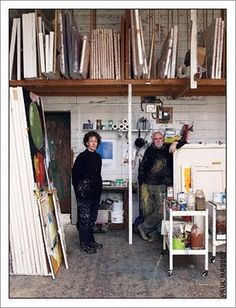 two people standing next to each other in front of bookshelves and shelves with paintings on them